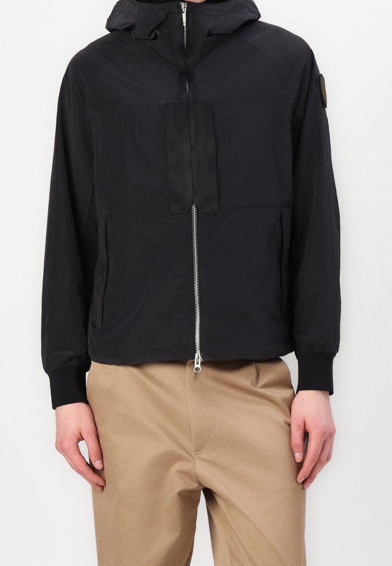 Veste noire à capuche avec fermeture éclair frontale et poignets côtelés portée avec un pantalon beige par une personne debout.