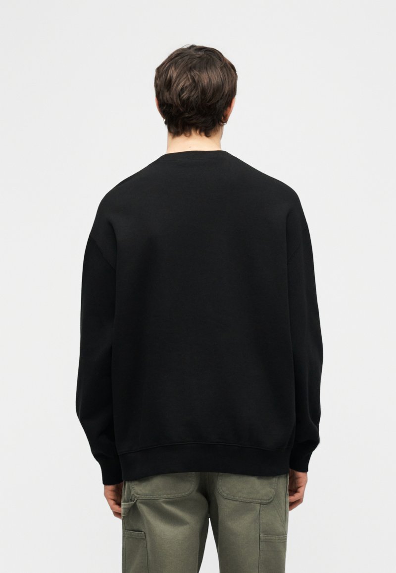 Uomo con capelli castani corti che indossa una felpa nera semplice e pantaloni cargo verde oliva, visto di spalle su sfondo bianco.