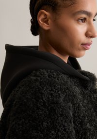 Zwart fleece jack met een gestructureerd, pluizig oppervlakte, met een zwarte stoffen capuchon en soepele kraag. Close-up van het profiel van een model.