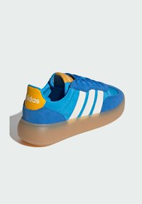 Plava tenisica od antilopa s bijelim kožnim prugama, narančastom petnom petljom i prozirnim potplatom. Zaobljen dizajn s teksturiranim tkaninom.