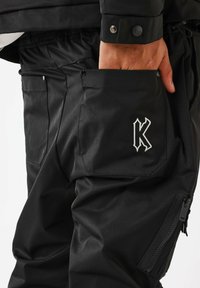 Pantaloni impermeabili neri con finitura strutturata, molteplici tasche e un distintivo logo bianco "K". Sono dotati di chiusure con cerniera e automatiche.