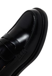 Mocasines de cuero negro con un acabado liso y pulido, que cuentan con una correa decorativa y detalles de costura a lo largo de los lados y la suela.