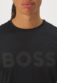 T-shirt de sport noir à manches courtes avec texture côtelée, affichant le mot "BOSS" en design rayé audacieux horizontalement sur la poitrine.