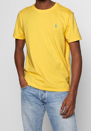 Geel katoenen T-shirt met ronde hals en korte mouwen. Bevat een klein blauw logo op de linkerkant van de borst. Gekleed met vervaagde blauwe jeans.