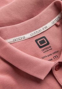 Rosa bomullspolo med strukturerad yta. Har en ribbad krage, logotags och en etikett som anger 100% bomull och storlek M.