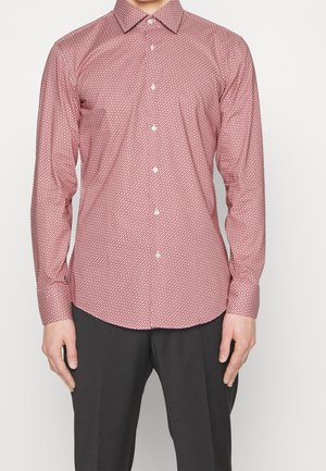 Mies, jolla on päällään pitkähihainen, napitettu punainen paita, jossa on pieni valkoinen geometrinen kuvio, ja tummanharmaat housut, seisoo yksiväristä taustaa vasten.