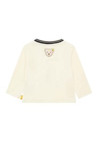 Langarm-Cremeshirt mit einem strukturierten Bärenpatch auf dem Rücken, schwarzem gestreiften Ausschnitt und einem kleinen gelben Etikett am Saum.