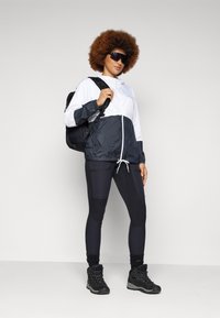 Weißer und marineblauer Windbreaker mit verstellbarem Kapuzen; schwarze Leggings mit Taschen; schwarzer Rucksack; robuste schwarze Wanderschuhe.