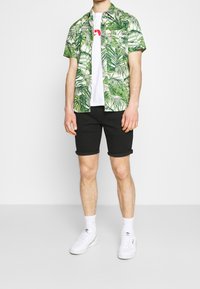 Hombre que lleva una camisa de manga corta blanca y verde con patrón de hojas sobre una camiseta gráfica blanca, pantalones cortos negros remangados, calcetines blancos y zapatillas Adidas blancas.
