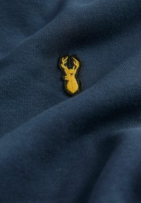 Tissu bleu marine avec une texture douce, présentant un petit logo de cerf brodé en jaune, souligné de noir. Le design est subtil et compact.