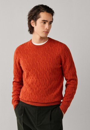Joven con pelo oscuro que lleva un suéter naranja de punto trenzado sobre una camisa blanca y pantalones negros, mirando hacia un lado con las manos en los bolsillos.