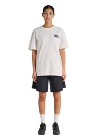 Kvinde stående iført en løs hvid T-shirt med logo, sorte knælange shorts, hvide sokker og sorte sneakers på en hvid baggrund.