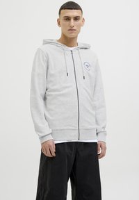 Sudadera gris con cremallera y capucha ajustable; presenta un logo circular azul en el lado izquierdo del pecho; lleva puesta una camiseta blanca y pantalones cortos negros.