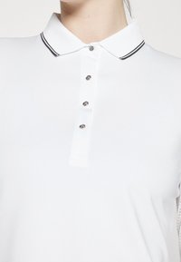 Biały polo z miękkiej tkaniny, z klasycznym kołnierzykiem w czarnym pasku, pięcioma guzikami z przodu oraz delikatnymi detalami tekstury.