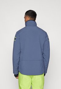 Chaqueta aislante azul con cuello alto, que presenta acentos en negro y verde neón en los hombros, y un ajuste relajado con una textura suave.