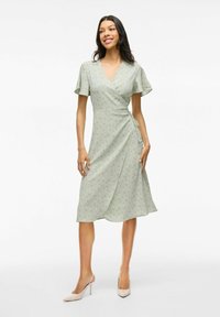 Robe portefeuille vert clair avec de courtes manches volantes, un motif floral et un détail de nœud sur le côté. Tissu lisse et coupe mi-longue.