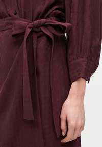 Μπορντό φόρεμα με διακριτικό γεωμετρικό σχέδιο, μακρύ φουσκωτό μανίκι με μανσέτα και κουμπί, και ζώνη δέσιμη στη μέση, που δείχνει ένα μέρος του χεριού.