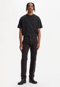 Hombre con cabello trenzado que lleva una camiseta negra, jeans oscuros y botas de cuero negras, de pie contra un fondo blanco.