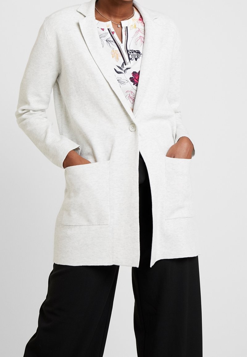 Femme portant un blazer gris clair à un bouton sur un chemisier à fleurs, avec un pantalon noir à jambes larges, mains dans les poches du blazer.