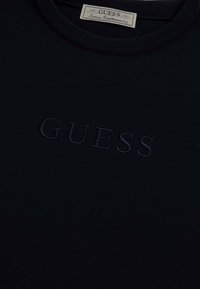 Sudadera azul marino con escote redondo, que presenta un logotipo "GUESS" en relieve en la parte delantera en un tono más oscuro. Textura de tela suave.