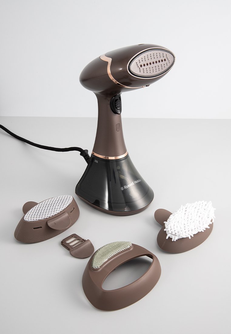 Steamer de prendas de mano marrón con cabezal redondeado, depósito de agua transparente y tres cepillos intercambiables en varias formas y texturas.