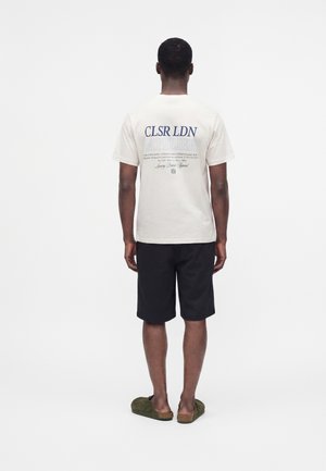 Uomo visto di spalle che indossa una maglietta bianca con grafica, pantaloncini neri e scarpe slip-on verdi su uno sfondo bianco semplice.