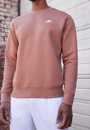 Person wearing a dusty pink Nike sweatshirt with white logo and white pants standing against a brick and light-colored wooden wall.