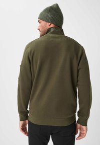 Felpa verde oliva con colletto alto, texture a coste e logo discreto sul retro. Abbinata a pantaloni neri slim.