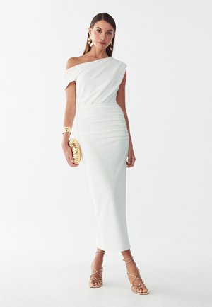 Frau trägt ein weißes One-Shoulder-Midikleid mit geraffter Seite, goldene Ohrringe, Armband, Clutch und Riemchen-Sandaletten mit hohen Absätzen vor weißem Hintergrund.