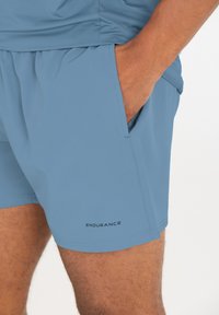 Lyseblå træningsshorts lavet af åndbart materiale, med elastisk talje, sidelommer og et lille "ENDURANCE" logo på kanten.