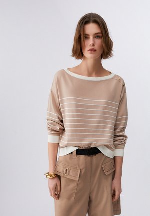 Donna con capelli corti castani che indossa una maglia a maniche lunghe beige e bianca a righe, pantaloni beige con tasche, cintura nera e braccialetti dorati al polso sinistro.