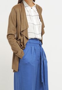 Cardigan marrone drappeggiato su una camicia bianca con motivo a griglia, abbinato a pantaloni blu a vita alta con cintura annodata e pieghe.
