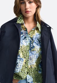 Navy jacket over a multicolored floral shirt with green, blue, and yellow patterns. Smooth texture, button-up design, and a collared neck.