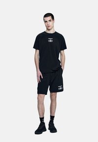 Conjunto atlético negro que incluye una camiseta de manga corta y pantalones cortos, ambos con logotipos blancos en contraste. El calzado son zapatillas negras con cordones.