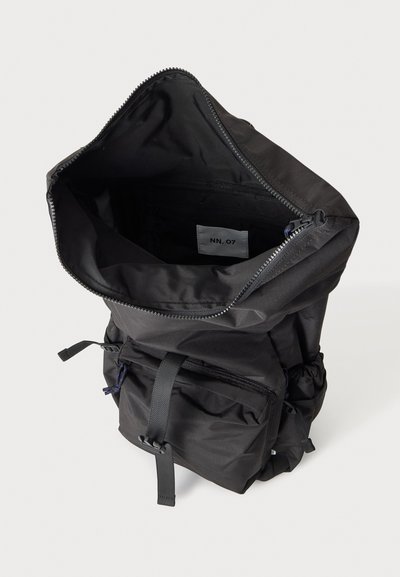 Sac à dos noir avec fermeture éclair supérieure ouverte révélant une petite étiquette à l'intérieur marquée "NN. 07,", poche avant avec fermeture par sangle à boucle visible.