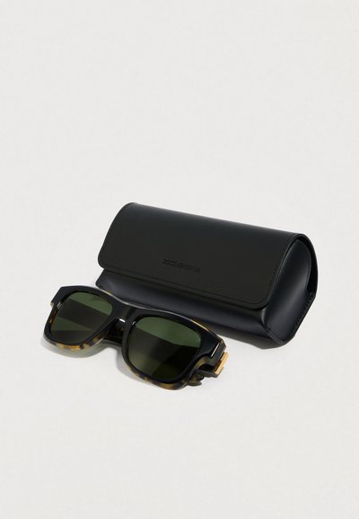 Črne sončna očala z vzorcem želvje lupine, temno zelenimi stekli in zlatimi poudarki, postavljena poleg črne usnjene etuije.
