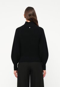 Schwarzer Strickpullover mit hohem Kragen und gerippter Textur. Mit Ballonärmeln und einem kleinen Logo auf dem oberen Rücken.