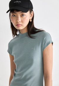 Šviesiai mėlyna trumpomis rankovėmis palaidinė su aukštu apykaklės kirpimu ir mažu siuvinėtu „CK“ logotipu ant krūtinės, derinama su juodu kepurėle.