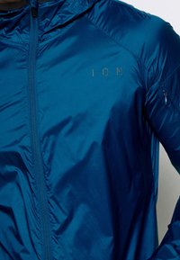 Närbild av en person som bär en glänsande blå zip-up jacka med en liten dragkedjeficka på ärmen och "ION" tryckt på bröstet.