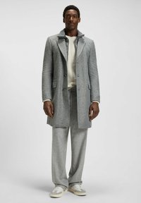 Grå rutiga kappa med notch-krage, tillsammans med en krämfärgad stickad tröja och matchande grå byxor. Skorna är ljusgrå sneakers.