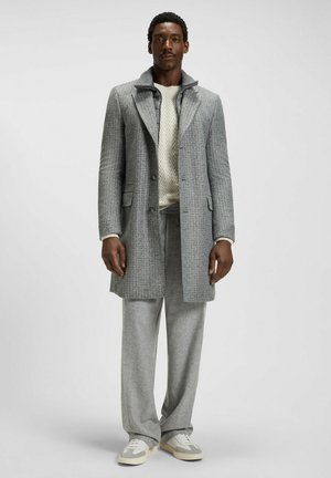 Manteau à carreaux gris avec col cranté, associé à un pull en tricot crème et un pantalon gris assorti. Les chaussures sont des baskets gris clair.