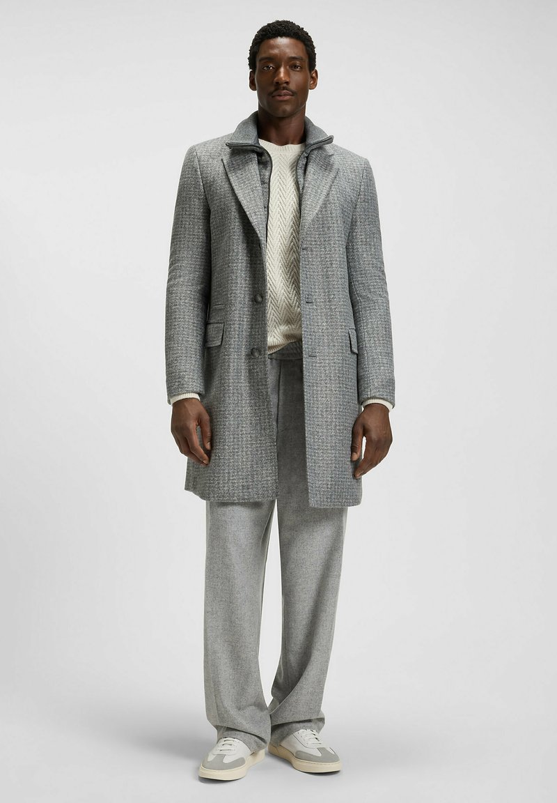 Grå rutiga kappa med notch-krage, tillsammans med en krämfärgad stickad tröja och matchande grå byxor. Skorna är ljusgrå sneakers.