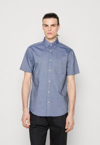 Bărbat cu părul pieptănat pe spate poartă o cămașă albastră cu mâneci scurte și buzunar la piept, asortată cu pantaloni negri, pe un fundal simplu.