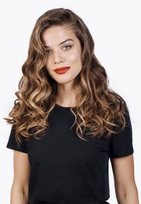 Portrait d'une personne avec de longs cheveux ondulés châtain clair et des yeux verts frappants. Elle porte une chemise noire et un rouge à lèvres rouge vif.