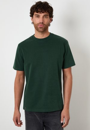 Homme aux cheveux bouclés foncés portant un t-shirt vert foncé à manches courtes et un jean foncé, debout devant un fond clair uni.