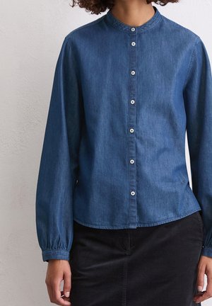 Kvinde iført en langærmet blå denim-skjorte med knapper og rund krave samt sort nederdel, stående mod en hvid væg.