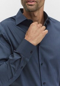 Granatowa koszula z długim rękawem, ozdobiona małym, powtarzającym się wzorem w kropki. Wykonana z gładkiego materiału, z usztywnionym kołnierzykiem i zapinanymi mankietami.