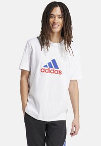 T-shirt bianca in cotone con un grande logo Adidas blu e rosso sulla parte anteriore. Vestibilità casual con maniche corte e scollatura rotonda.