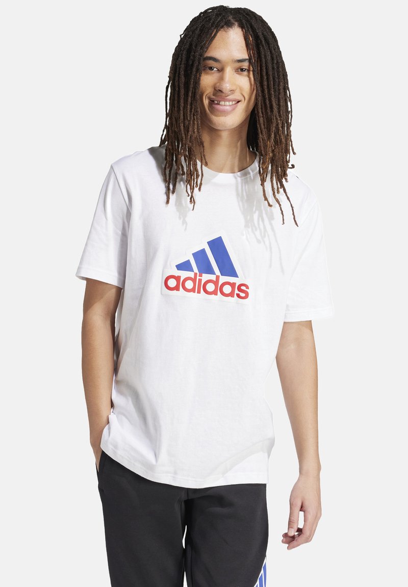 T-shirt bianca in cotone con un grande logo Adidas blu e rosso sulla parte anteriore. Vestibilità casual con maniche corte e scollatura rotonda.