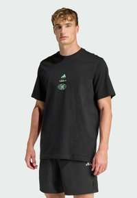 Camiseta de algodón negra con mangas cortas, que presenta un logotipo de Adidas en verde y un gráfico de texto en el centro. Diseño simple y de corte relajado.